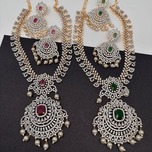 May include: Two ornate necklace and earring sets featuring gold-tone chains, intricate pavé-set clear stones, and dangling white pearls. One set features deep red rectangular center stones, while the other set features deep green rectangular center stones.