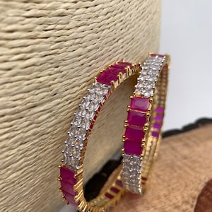 May include: Two gold bangles, each featuring alternating rows of bright pink and clear rectangular stones. The jewellery is displayed against a neutral backdrop, highlighting the bangles' design and sparkle.