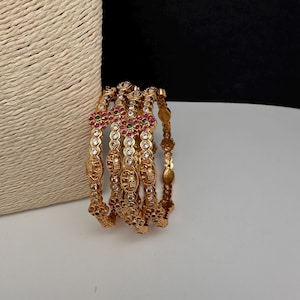 May include: A set of four gold-toned bangles featuring intricate metalwork, embedded with clear white stones, small red floral accents, and tiny green center stones. The bangles display a repeating pattern of circular and oval shapes with detailed engravings.