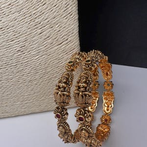 May include: Gold-colored bangles with intricate carvings of figures and floral designs. The bangles feature red and green gemstone accents. The jewelry is displayed against a neutral background.