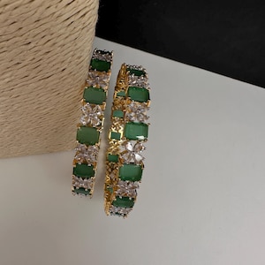 May include: Two gold-toned bangles with emerald green and clear crystal accents. The bangles feature rectangular green stones and clear crystal flower-shaped designs. The jewelry is displayed on a white surface.