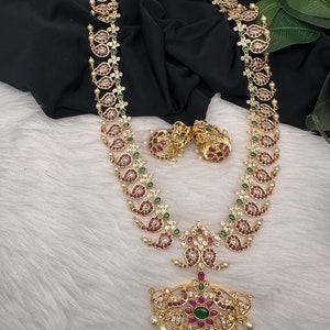 May include: Gold-toned necklace and earring set featuring a paisley design with red, green, and clear gemstones. The necklace has a large pendant with a central green stone and dangling pearls. The earrings mirror the necklace's design.