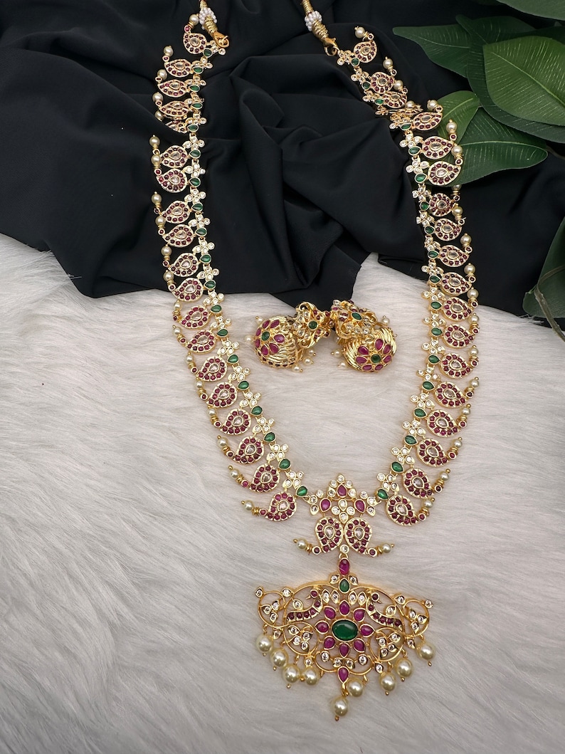May include: Gold-toned necklace and earring set featuring a paisley design with red, green, and clear gemstones. The necklace has a large pendant with a central green stone and dangling pearls. The earrings mirror the necklace's design.