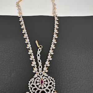 May include: An ornate gold-toned headpiece with clear and ruby-colored gemstones and faux pearls. The headpiece features a central circular design with a ruby-colored stone, surrounded by clear gemstones and dangling pearls.