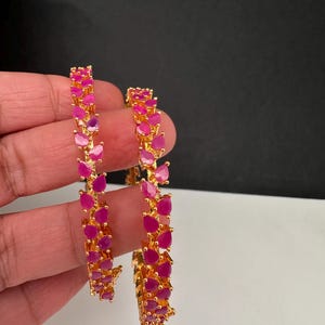 Puede incluir: Dos brazaletes dorados con una hilera continua de piedras de color rosa vibrante en forma de l&aacute;grima. El engaste de metal pulido sujeta cada piedra con garras, creando un patr&oacute;n texturizado y repetitivo a lo largo de la banda circular.