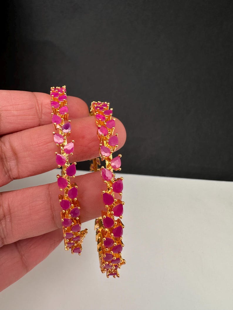 Puede incluir: Dos brazaletes dorados con una hilera continua de piedras de color rosa vibrante en forma de l&aacute;grima. El engaste de metal pulido sujeta cada piedra con garras, creando un patr&oacute;n texturizado y repetitivo a lo largo de la banda circular.