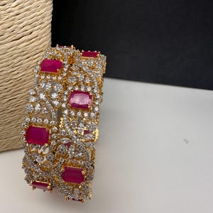 May include: Gold-toned bangles with a floral design, featuring rectangular ruby-red gemstones and clear, sparkling crystals. The intricate detailing and vibrant colors create a luxurious look. These bangles are a statement piece for any jewelry collection.