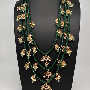 May include: A three-strand emerald green beaded necklace with gold-toned pendants and accents. The pendants have red and white accents. The necklace is displayed on a black mannequin.