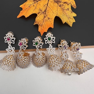 May include: Six pairs of gold-toned, ornate earrings with various colored gemstones. Each earring features a floral design at the top and a bell-shaped bottom, encrusted with small, clear stones. A large, yellow leaf is in the background.
