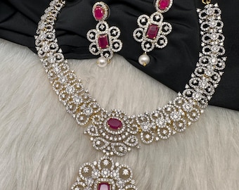 Puede incluir: Un deslumbrante conjunto de joyas con collar y pendientes a juego. El collar y los pendientes están adornados con piedras claras y brillantes y gemas rojas vibrantes. El collar tiene un colgante central con una gran piedra roja y una perla colgante.
