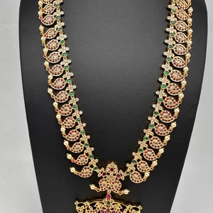 May include: A gold-toned necklace with a paisley design, featuring red, green, and clear gemstones, and small white pearls. The necklace is displayed on a black stand, showcasing its intricate details and craftsmanship.