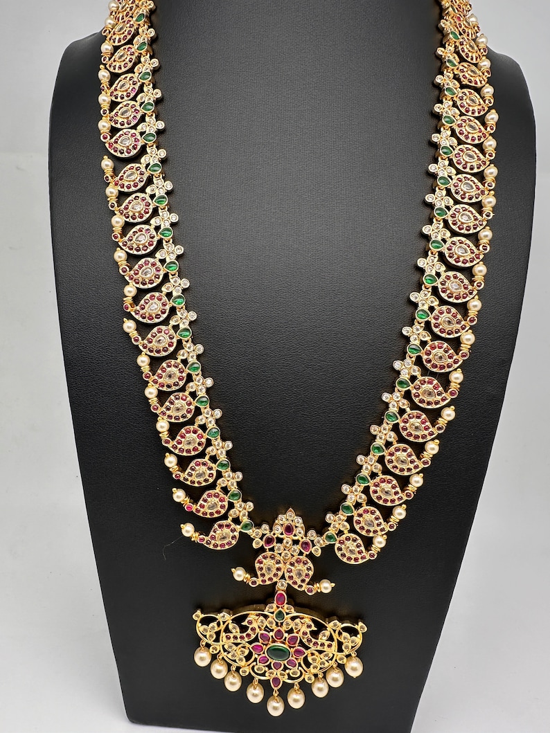 May include: A gold-toned necklace with a paisley design, featuring red, green, and clear gemstones, and small white pearls. The necklace is displayed on a black stand, showcasing its intricate details and craftsmanship.