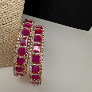 May include: Two gold-toned bangles with alternating fuchsia-colored square stones and small, clear, round stones. The bangles are displayed on a white surface with a woven beige background.