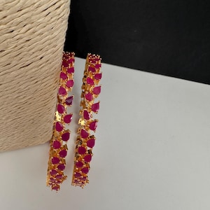 May include: Two gold-tone bangles featuring a continuous row of teardrop-shaped, vibrant fuchsia-pink stones. The metal setting is polished gold, and each bangle is constructed with a series of connected, prong-set stones in a uniform pattern.