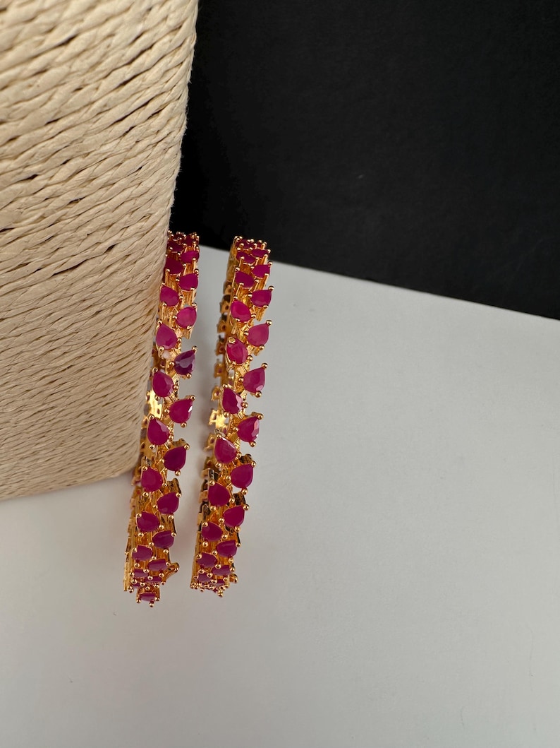 Puede incluir: Dos brazaletes dorados con una hilera continua de piedras en forma de l&aacute;grima de color rosa fucsia vibrante. El engaste met&aacute;lico es de oro pulido y cada brazalete est&aacute; construido con una serie de piedras engarzadas en un patr&oacute;n uniforme.