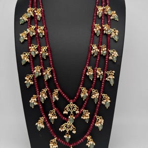 May include: A multi-strand necklace featuring deep red beads and gold-toned pendants. The pendants are adorned with green gemstones and small white pearls. The necklace is displayed on a black mannequin.