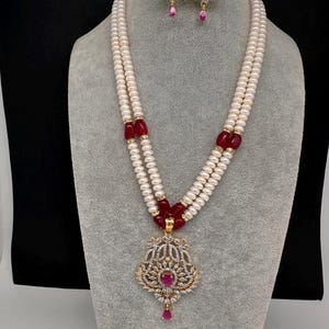 May include: A two-strand pearl necklace with a pendant and matching earrings. The pendant is gold-toned with clear and red gemstones. The necklace has red gemstone accents. The earrings have a similar design with a red teardrop gem.