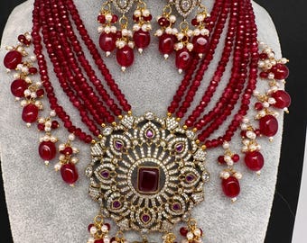 May include: A luxurious jewelry set featuring a necklace and matching earrings. The necklace has multiple strands of red beads, a large central pendant with a square red stone, and ornate gold-colored detailing. The earrings mirror the pendant design, with dangling red beads and white pearls.