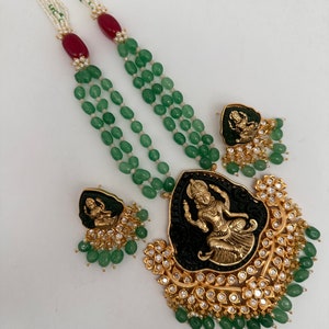 May include: An ornate necklace and earring set featuring a gold pendant with a black center and a gold figure. The necklace has multiple strands of green beads, white beads, and red accents. The earrings mirror the pendant design.