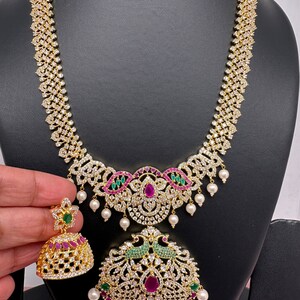 Op de afbeelding: Een goudkleurige ketting- en oorbellenset. De ketting heeft een diamantachtig patroon met een hanger versierd met rode en groene edelstenen en witte parels. De bijpassende oorbellen hebben een vergelijkbaar ontwerp. De sieraden worden gepresenteerd op een zwarte mannequin.