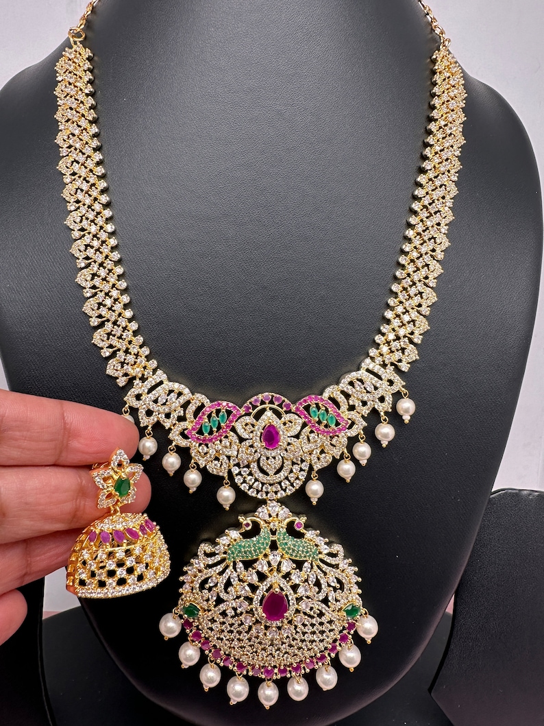 Op de afbeelding: Een goudkleurige ketting- en oorbellenset. De ketting heeft een diamantachtig patroon met een hanger versierd met rode en groene edelstenen en witte parels. De bijpassende oorbellen hebben een vergelijkbaar ontwerp. De sieraden worden gepresenteerd op een zwarte mannequin.