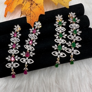 May include: Two pairs of dangling earrings. One pair features ruby-colored teardrop and oval-shaped stones, accented with clear crystals. The other pair has emerald-colored teardrop and oval-shaped stones, also with clear crystals. Both pairs have gold-colored metal.