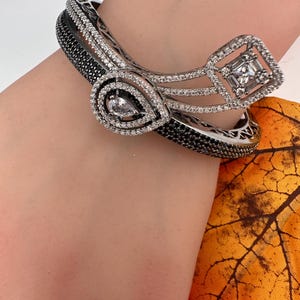 May include: A silver-toned bracelet with black and white accents, featuring a teardrop-shaped design and a square-shaped design, both adorned with clear gemstones. The bracelet is worn on a wrist.
