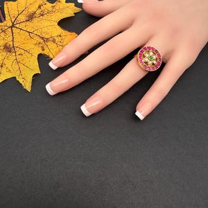 May include: A gold ring featuring a circular centerpiece with a border of square-cut pink stones surrounding a central flower motif. The flower has white petal-shaped stones and a small green stone at its center, set against a dark background.