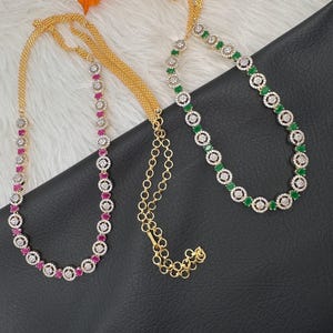 May include: Three gold-toned necklaces with gemstone accents. One necklace features alternating red gemstones and clear crystals, while another has green gemstones and clear crystals. The necklaces are displayed on a black surface.