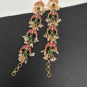May include: A pair of long gold-tone earrings featuring four stacked crescent-shaped segments. Each segment is inlaid with a pink stone and a central teardrop-shaped green stone, accented with small white pearl-like beads and a dangling chain link.