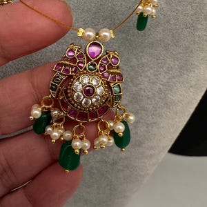 May include: A gold-toned necklace with a pendant featuring a central floral design surrounded by red and clear gemstones. The pendant is adorned with green teardrop-shaped beads and small white pearls. The necklace is held by a hand.