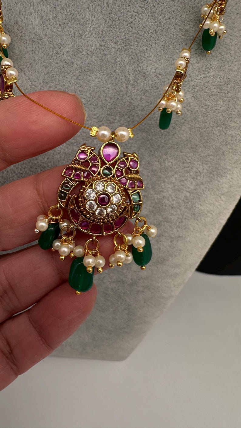 May include: A gold-toned necklace with a pendant featuring a central floral design surrounded by red and clear gemstones. The pendant is adorned with green teardrop-shaped beads and small white pearls. The necklace is held by a hand.