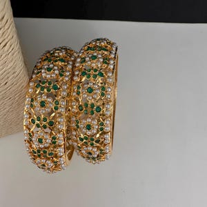 May include: Two gold bangles with intricate floral patterns. The bangles are adorned with green and white gemstones, creating a luxurious and ornate design. The jewelry is displayed on a white surface with a neutral background.