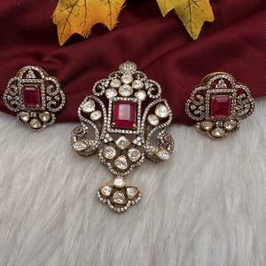 May include: A jewelry set consisting of a pendant and two matching earrings. Each piece features a central rectangular red stone surrounded by smaller white stones set in an intricate, dark metal frame with a filigree-like, ornate design.