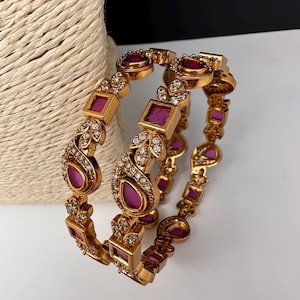 May include: Gold-toned bangles with square and teardrop-shaped magenta gemstones and small clear stones. The bangles have an ornate design with floral and geometric elements. The jewelry is displayed on a white surface.