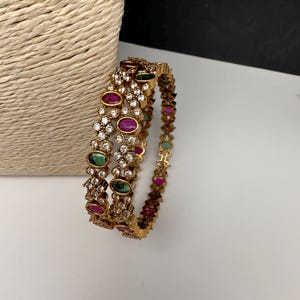 May include: Gold-toned bangles with embedded gemstones. The bangles feature a repeating pattern of clear crystals, oval-shaped green and red gemstones. The jewelry is displayed on a white surface.