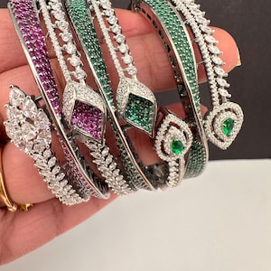 May include: A collection of silver-toned bangles, each adorned with vibrant gemstones. The bangles feature alternating rows of sparkling white stones and either ruby-red or emerald-green gems. The designs include leaf and teardrop shapes.