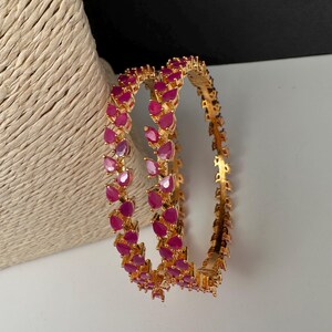 Puede incluir: Un par de brazaletes dorados con una hilera continua de piedras de color rosa vibrante en forma de l&aacute;grima. La banda de metal tiene un acabado pulido y las piedras est&aacute;n engastadas en un patr&oacute;n repetitivo alrededor de toda la circunferencia.