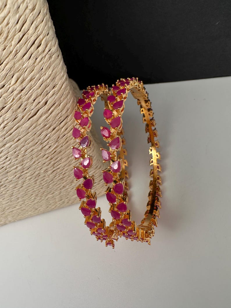 Puede incluir: Un par de brazaletes dorados con una hilera continua de piedras de color rosa vibrante en forma de l&aacute;grima. La banda de metal tiene un acabado pulido y las piedras est&aacute;n engastadas en un patr&oacute;n repetitivo alrededor de toda la circunferencia.