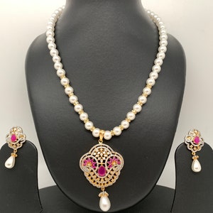 May include: A pearl necklace and matching earrings set. The necklace features a strand of white pearls with gold-colored accents. The pendant and earrings have a gold-colored filigree design with red and clear gemstones, and a dangling white teardrop pearl.