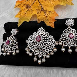 May include: A jewelry set featuring a pendant and matching earrings. The pendant and earrings have a floral design with clear stones and a central red stone. The earrings and pendant are adorned with dangling pearls. A yellow leaf is in the background.