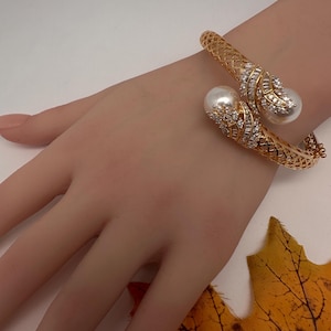 May include: Gold-toned cuff bracelet featuring a filigree design. The bracelet has two large, white, pearl-like accents, each surrounded by sparkling, clear crystals. A luxurious piece of jewelry.