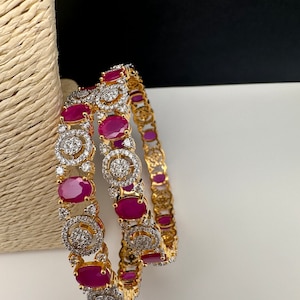 May include: Two gold bangles with oval, ruby-colored stones and clear, round accent stones. The bangles have a repeating pattern of stones and are displayed on a white surface with a neutral background.