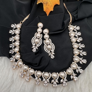 May include: A gold-toned necklace and matching earrings set featuring large, round, faux pearls and clear, sparkling crystals. The necklace and earrings are arranged on a black fabric background, with a yellow leaf in the upper corner.