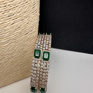 May include: A set of two gold-toned bangles. Each bangle is encrusted with small, clear crystals and features two large, emerald-green, rectangular stones. The bangles are displayed against a neutral background.