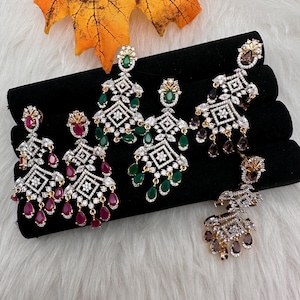 May include: An assortment of ornate chandelier earrings with various gemstone colors, including ruby red, emerald green, and amethyst purple. The earrings feature a geometric design with clear, sparkling accents. Displayed on a black velvet surface.