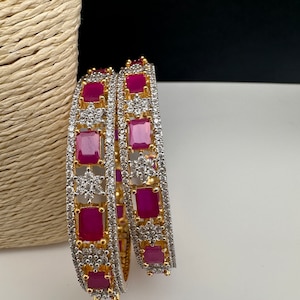 May include: Two gold and silver bangles with rectangular ruby-red stones and small clear stones. The bangles are set with a gold band and a silver band, and the ruby-red stones are evenly spaced around the circumference.