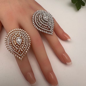 May include: Two large, ornate rings with a teardrop design. One ring is gold-toned, the other is silver-toned, both encrusted with numerous small, sparkling stones. The rings are displayed on a hand with manicured nails.