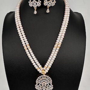 May include: A pearl necklace and earring set. The necklace features two strands of white pearls with gold accents and a decorative pendant. The earrings and pendant have a floral design with clear gemstones and a teardrop-shaped dangle.