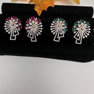 May include: Four pairs of stud earrings with a floral design. Two pairs feature ruby-colored stones, and the other two have emerald-colored stones. Each earring has a gold-colored center and is surrounded by clear, sparkling stones.